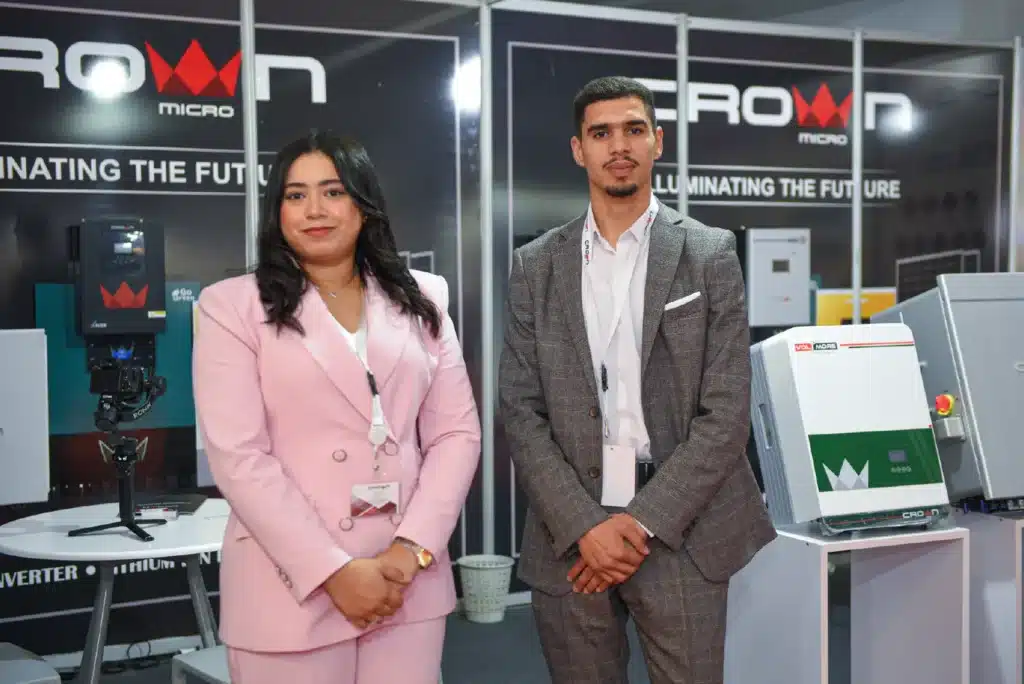 A woman in a pink suit and a man in a grey blazer standing by solar inverter displays at a Crown Micro booth.