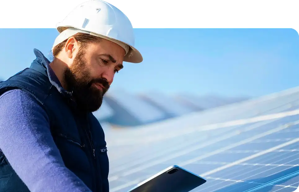 Male technician in white hard hat and blue vest using a tablet at a large-scale solar panel installation.